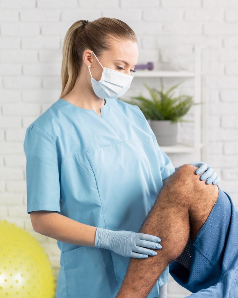 A masked medical professional in scrubs carefully examining a patient's knee prior to a joint treatment.