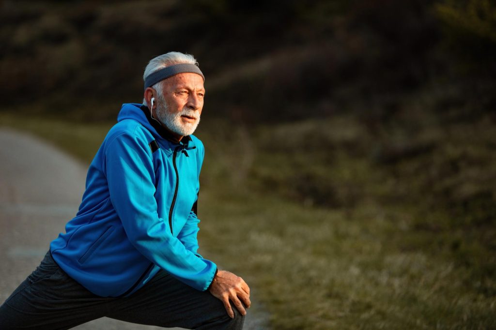 An active senior man with a white beard wearing a blue athletic jacket and headband, stretching his legs on an outdoor path at sunset.