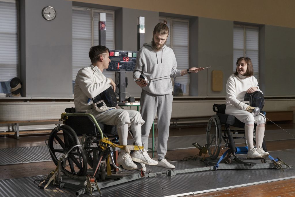 Two wheelchair fencers in protective white gear discussing technique with a coach using specialized fencing equipment in a gym.