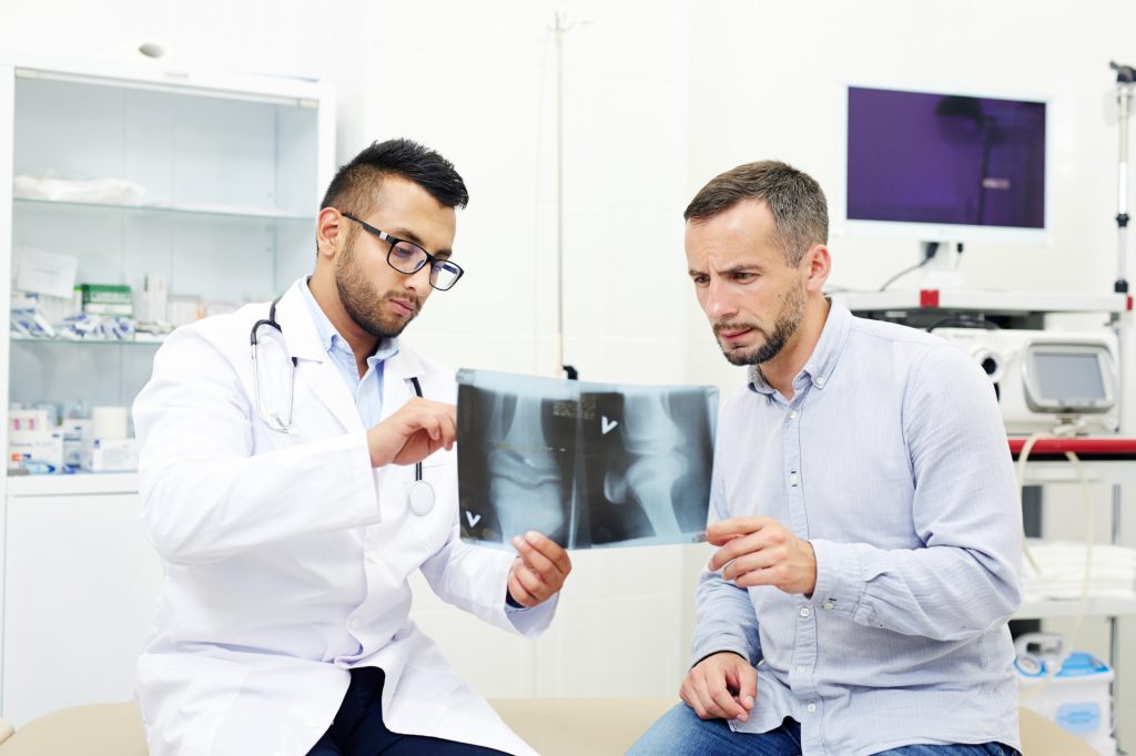 A male doctor in a white lab coat showing a physical knee X-ray printout to a concerned male patient in a medical office.