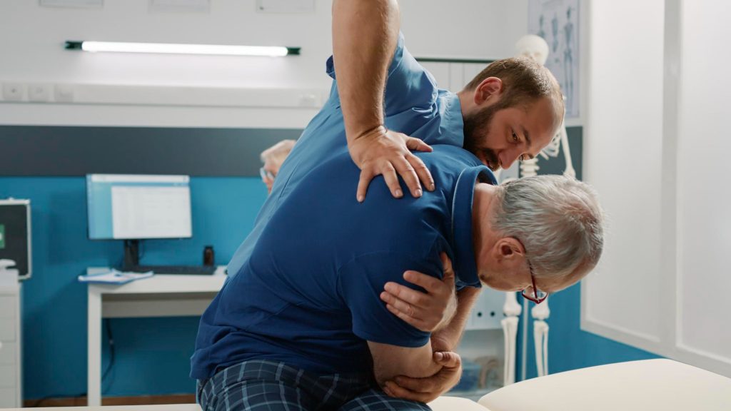 A physical therapist assisting a senior male patient with a seated back and shoulder stretch in a clinic.