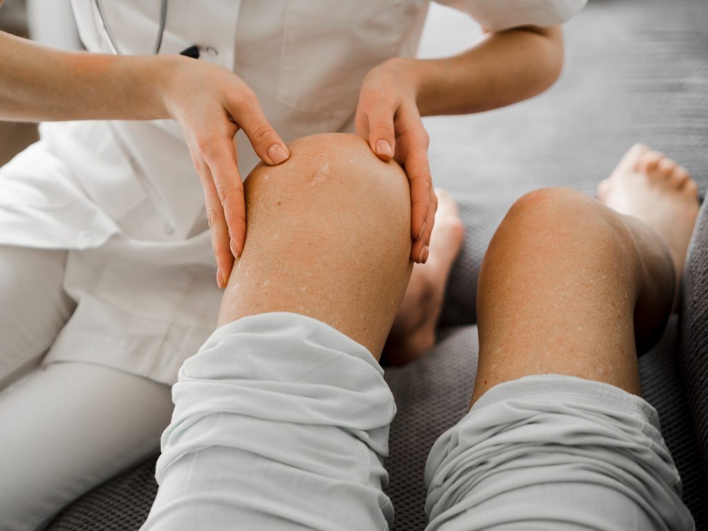 A close-up of a medical professional's hands gently examining a patient's bare knee during an orthopedic consultation.