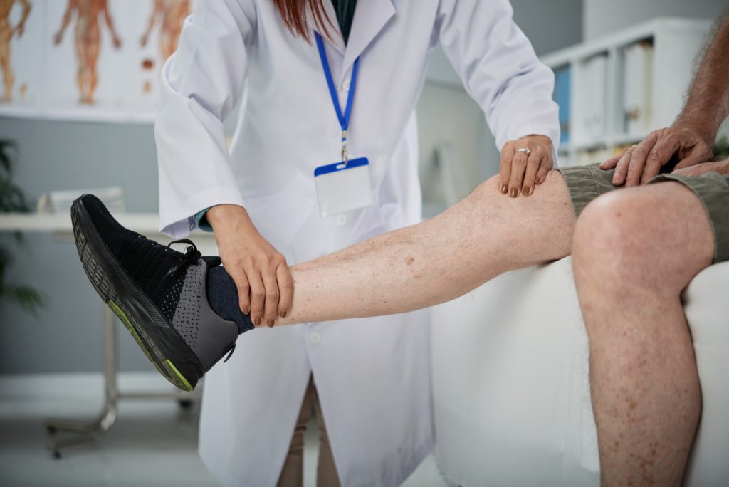 A doctor in a white lab coat examining a male patient's leg and knee mobility in a bright medical clinic.