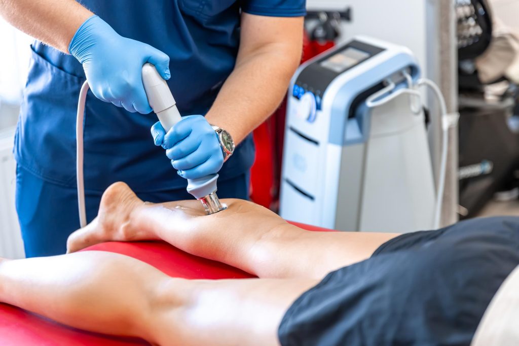 A medical professional in blue scrubs and gloves applying a shockwave therapy device to a patient's bare calf in a bright clinic setting.