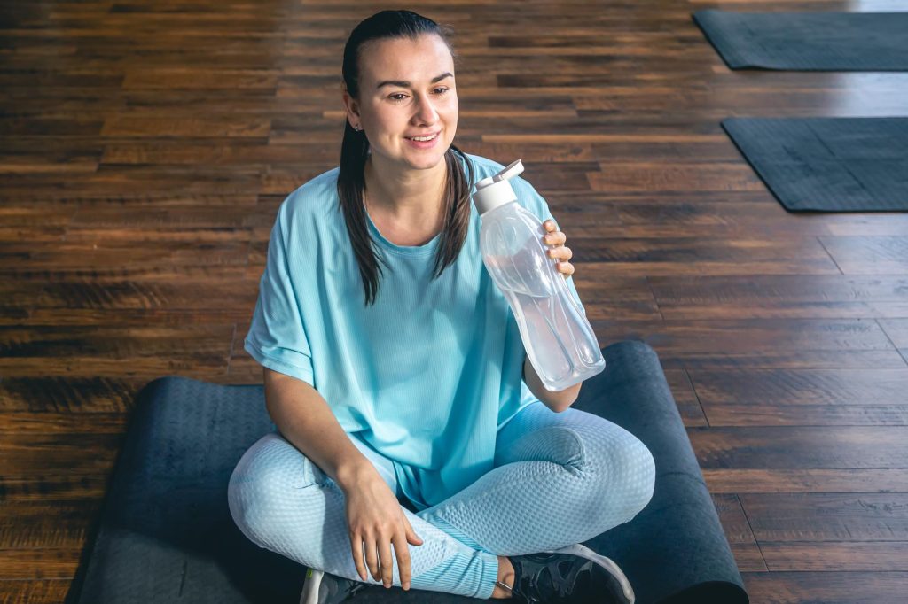 A woman in blue athletic wear sitting cross-legged on a black yoga mat in a gym, holding a clear water bottle.