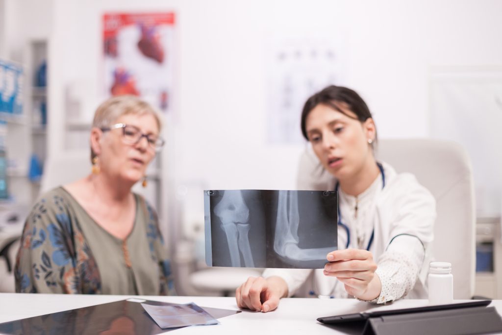 A female doctor in a white coat holding up an X-ray of a knee joint while discussing surgical options with a senior female patient in a hospital office.
